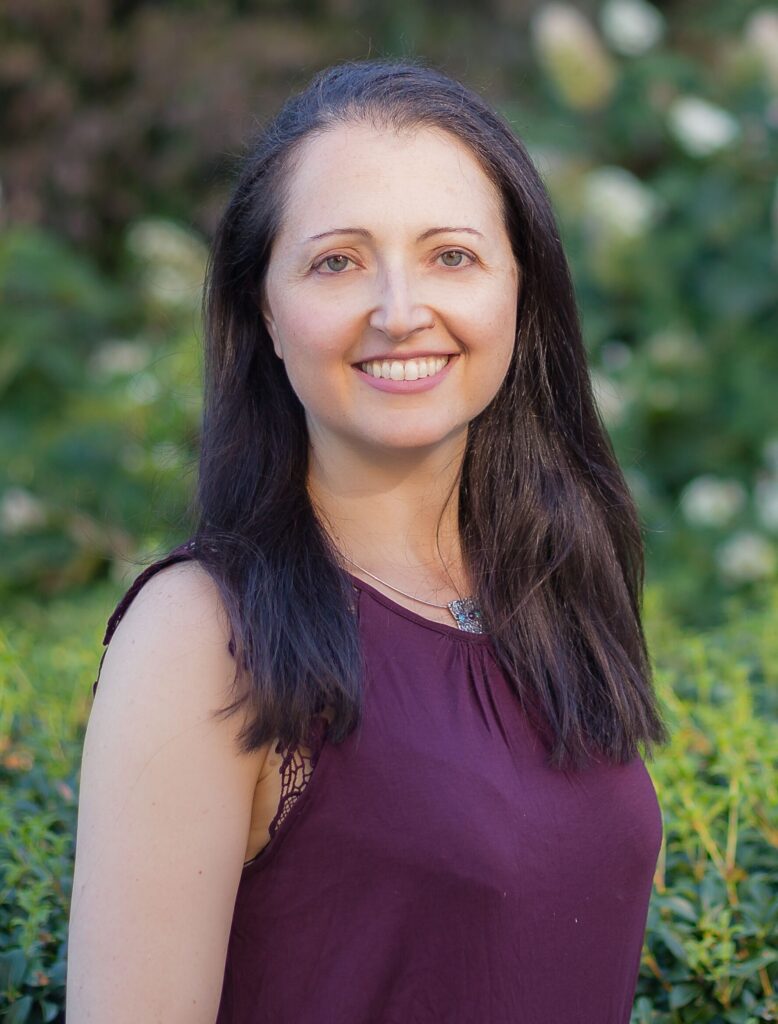 Headshot of a white middle aged woman with dark mid length hair and a burgundy sleeveless top. In the background are lush greenery and old university buildings.