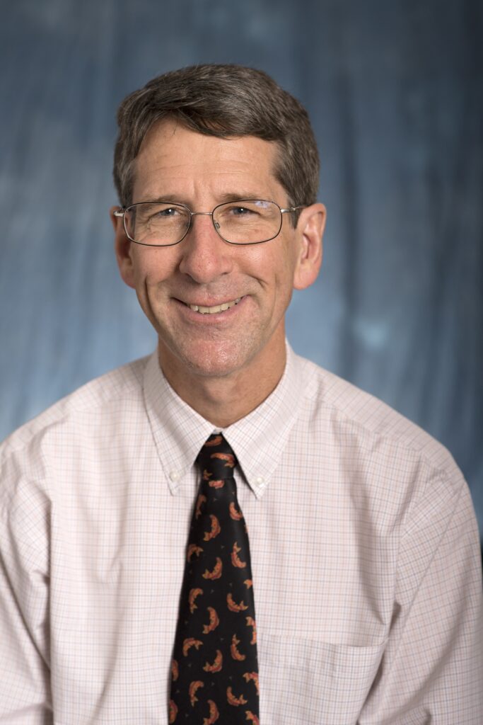 Headshot of a white middle-aged man with brown and grey hair and glasses wearing a tie.