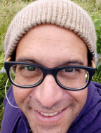 Close up facial image of a smiling middle aged white male wearing dark rimmed glasses and a light brown beanie, outdoors.