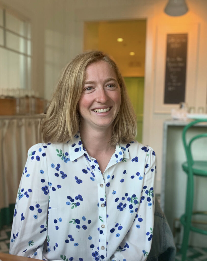 A white woman with blonde, shoulder length hair wearing a white button up with blueberries on it. In the background is a café.