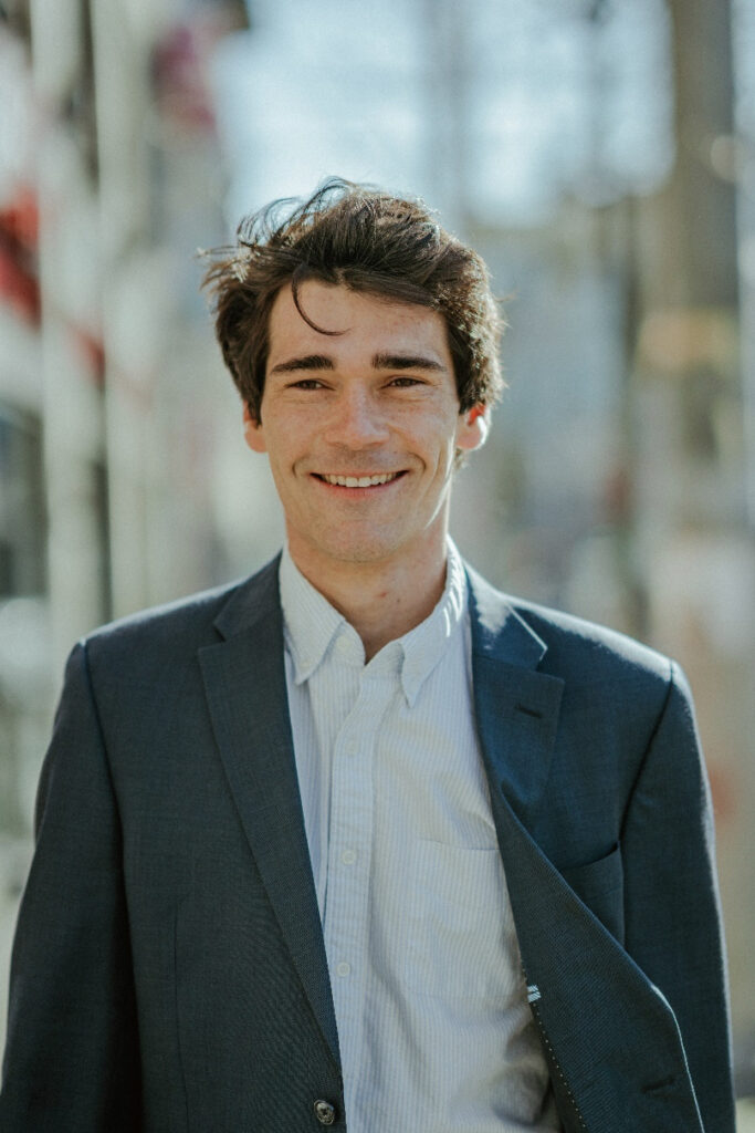 Headshot of a white young adult male with dark hair, wearing a light blue and white stripped button-down shirt and dark blue jacket. In the background, there are greyish buildings.