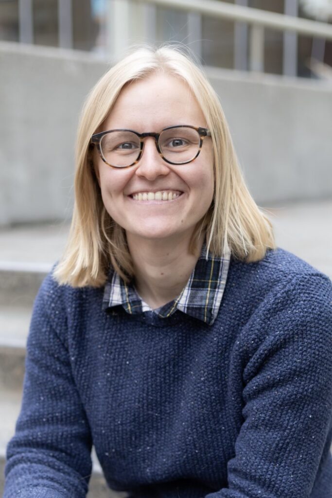 Headshot of a white woman mid-20s with short blonde hair and blue sweater sitting on concrete steps.