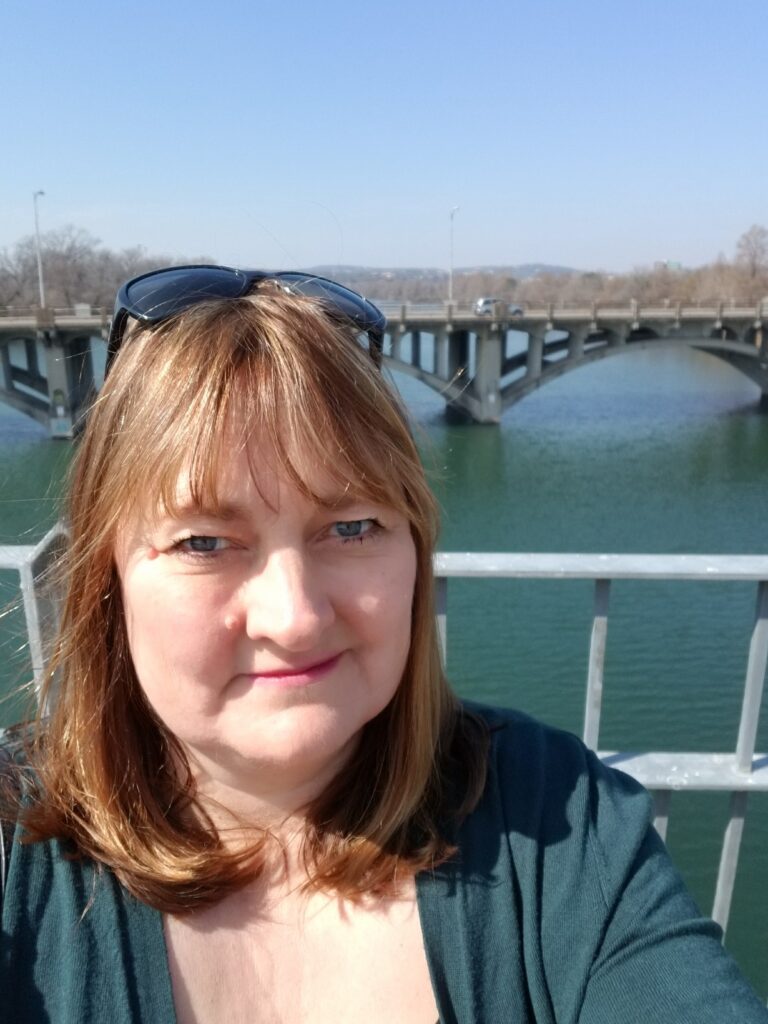 Picture of a white female with brown shoulder-length hair, blue yes, a smile and sunglasses on her head. In the background is a pine-green river, which matches her shirt, a bridge and trees in the distance.