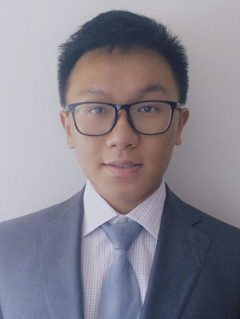 Headshot of a man with short hair, wearing black frame glasses and a gray business suit in plain background.