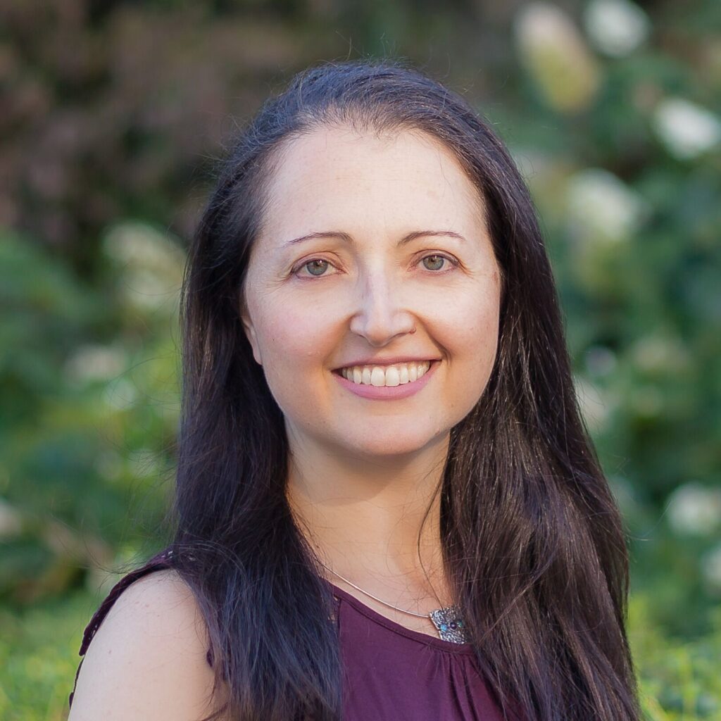 Headshot of a white middle aged woman with dark mid length hair and a burgundy sleeveless top. In the background is lush greenery.